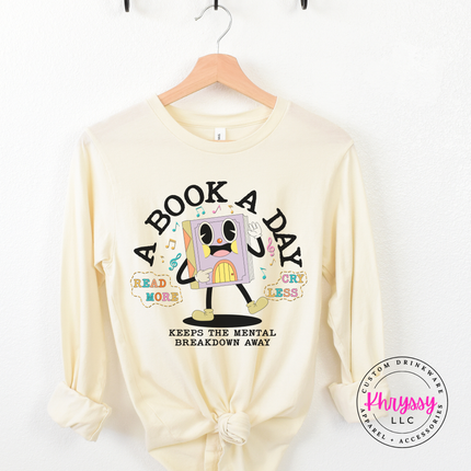Natural long sleeve showing the multicolor retro book character with read more cry less lettering.