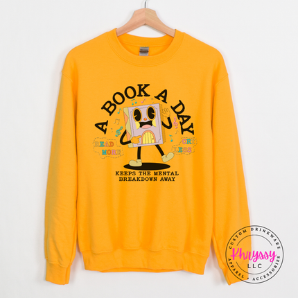 Gold crewneck sweatshirt featuring the pastel lavender book character and A Book A Day lettering.