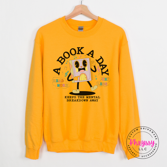 Gold crewneck sweatshirt featuring the pastel lavender book character and A Book A Day lettering.