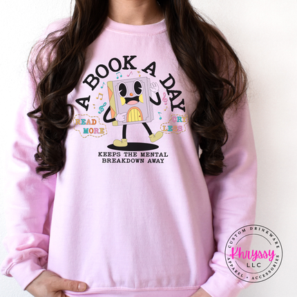 Light Pink Gildan crewneck printed with the retro cartoon A Book A Day Keeps The Mental Breakdown Away graphic.