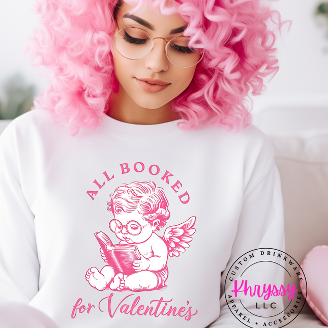 A woman with pink hair and glasses, sitting, wearing a white unisex sweatshirt with a graphic of a cherubic angel reading, and text that says 'All Booked for Valentine's Day'.