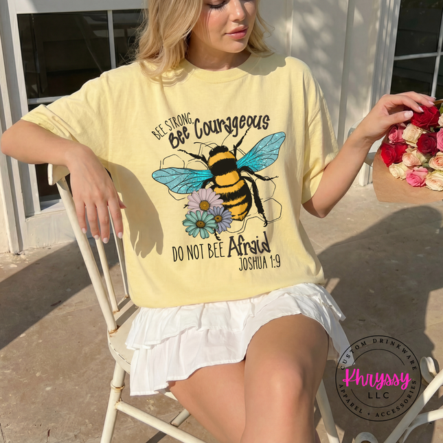 Woman wearing a yellow t-shirt with a bee graphic and text, sitting outdoors.
