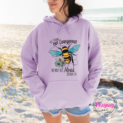 Person wearing a lavender hoodie with a bee design and text on a beach.