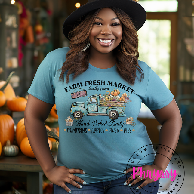 A person wearing a blue unisex shirt with a graphic print of a rustic truck and fall harvest market scene, standing in a store with pumpkins and apples displayed in the background.