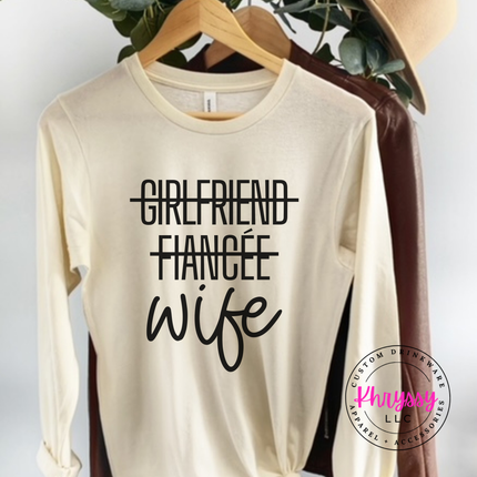 Beige long-sleeve shirt with 'Girlfriend', 'Fiancée', and 'Wife' text on a hanger.