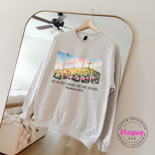 Ash crewneck sweatshirt with a He Has Risen watercolor cross and wildflowers graphic.