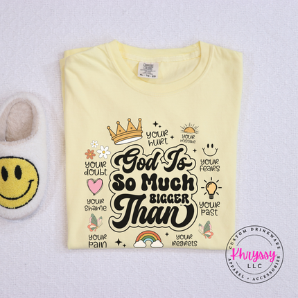 Butter garment-dyed tee with a God Is So Much Bigger Than faith graphic and icons.
