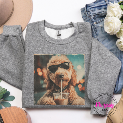 Gray sweatshirt with a dog wearing sunglasses and a drink graphic, placed on a surface with jeans and flowers.