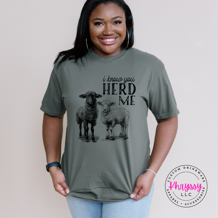 Woman wearing a moss colored t-shirt with sheep graphic and text, standing against a plain background.