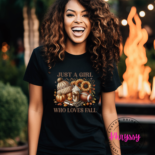 A woman smiling and wearing a black unisex t-shirt with a fall-themed graphic that includes the text 'Just a girl who loves fall' and depicts pumpkins and autumn leaves.