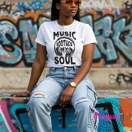 Woman wearing a white t-shirt with 'Music Soothes My Soul' text and graphics, sitting against a graffiti-covered wall.