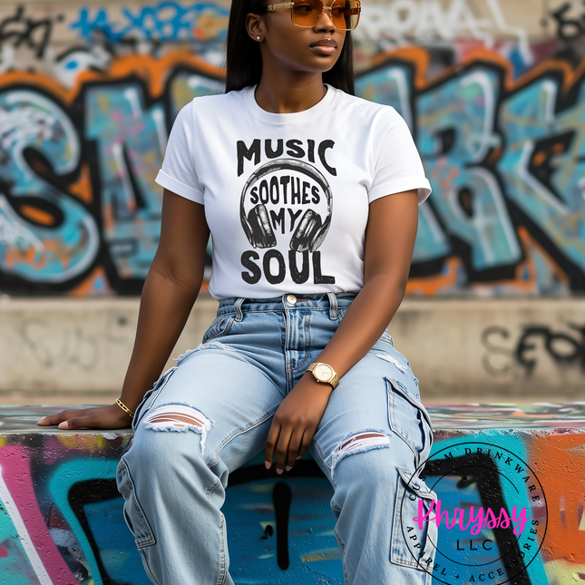 Woman wearing a white t-shirt with 'Music Soothes My Soul' text and graphics, sitting against a graffiti-covered wall.