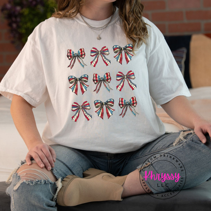 White garment-dyed tee displaying a patriotic coquette flag-bow graphic.