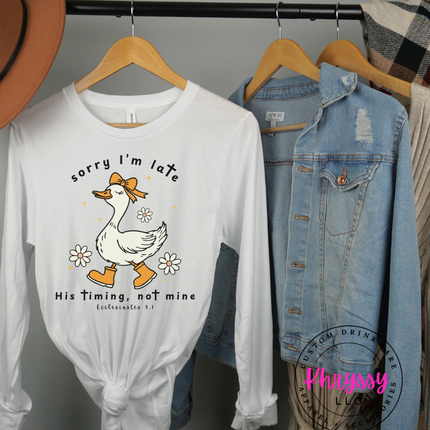 White long-sleeve shirt with a duck graphic and text on a rack with jackets and hats.