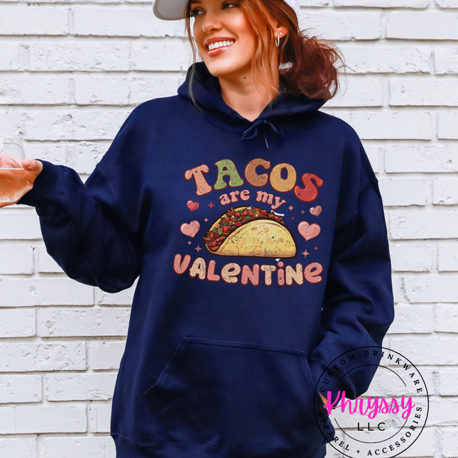 Person wearing a navy hoodie with 'Tacos are my Valentine' text and graphics against a white brick wall.