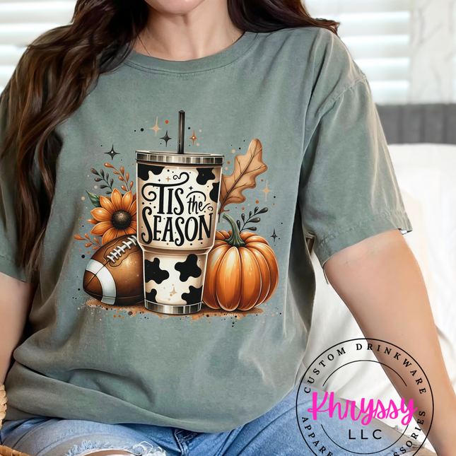 A woman smiling and wearing a long-sleeved unisex shirt with a fall-themed design, featuring pumpkins, a football, and autumn leaves.